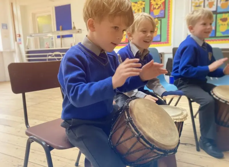 Three primary pupils in blue school jumpers seated on chairs in a school hall, each with a djembe drum held between their knees, striking the skins with both hands and grinning.