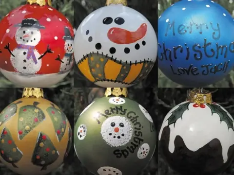 Six finished hand-painted baubles shown together: snowmen on red and white, a scarecrow face, Christmas trees on gold, a green bauble reading Merry Christmas, and a Christmas pudding with holly.