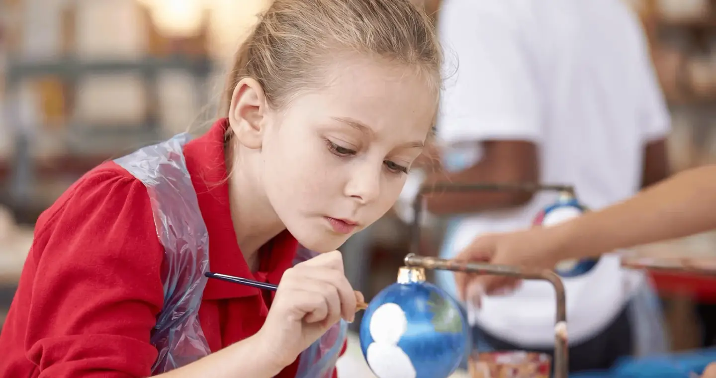 A pupil in a red school jumper and a plastic apron leaning in with a fine brush to paint a white snowman onto a blue bauble held upright on its stand, another child painting at the table behind her.