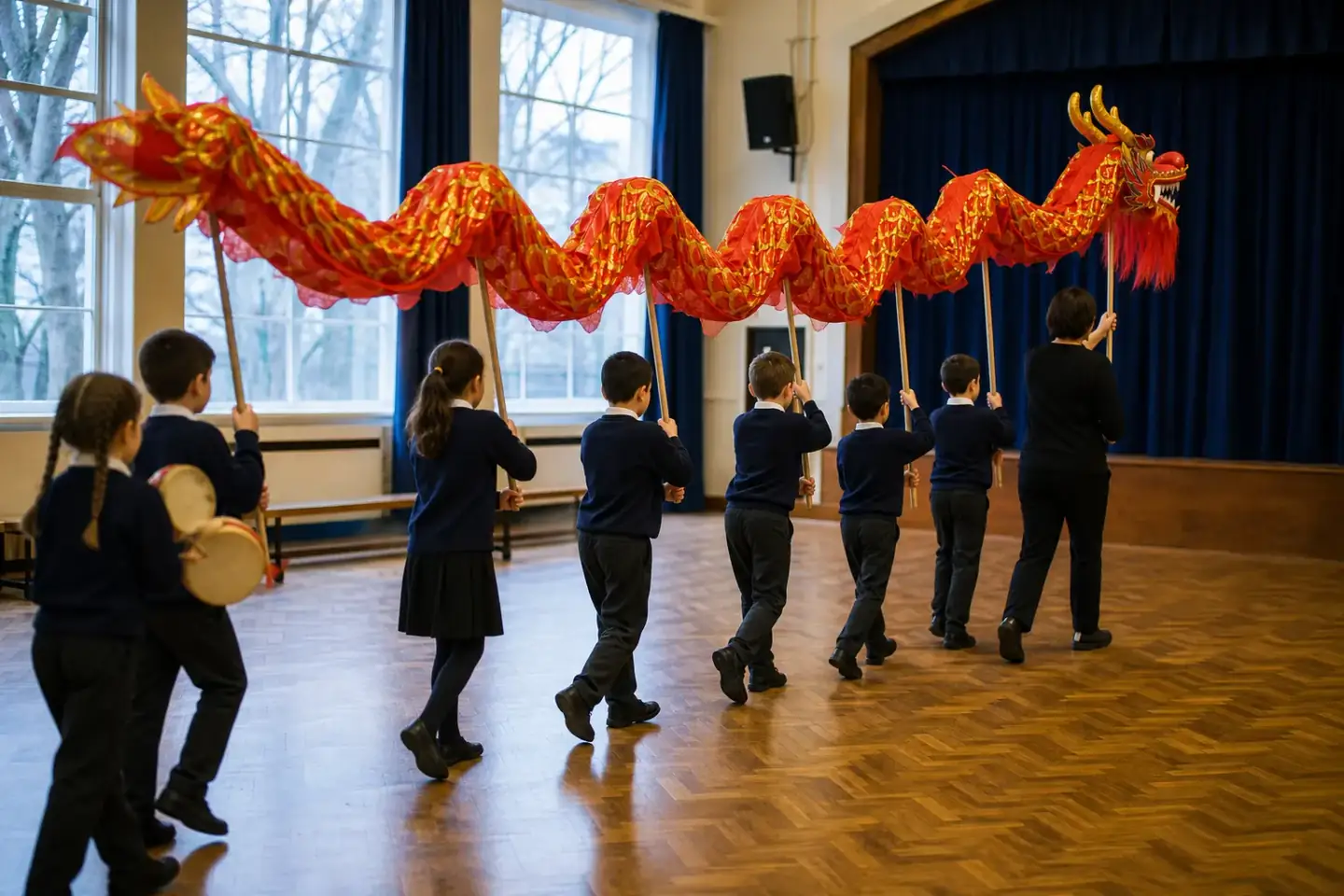 Pupils in school jumpers moving down a school hall carrying a long red and gold cloth dragon on wooden poles, seen from behind as the body ripples above their heads.