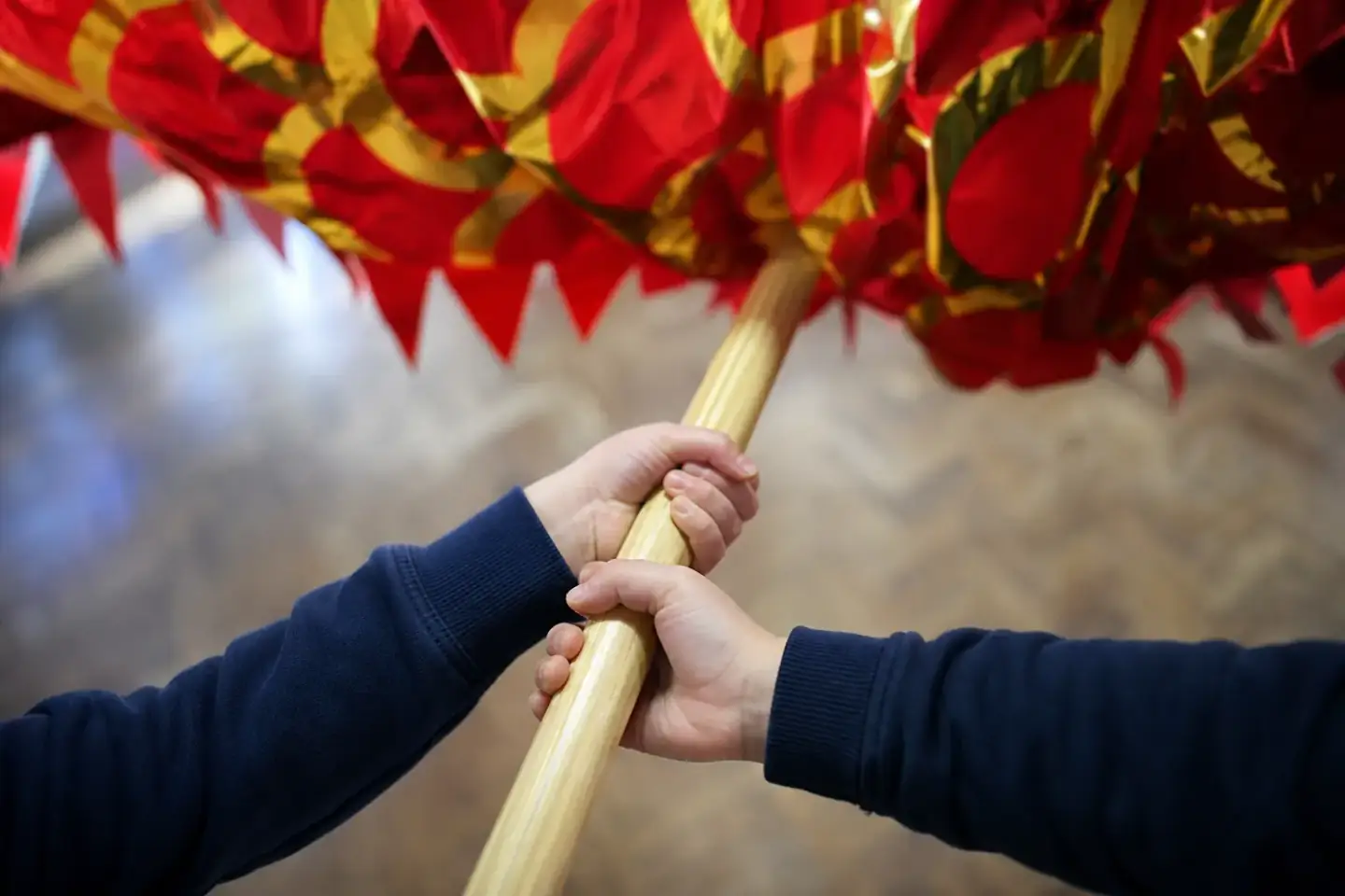 Close view of two pupils' hands gripping the varnished pole beneath a red and gold dragon segment, the cane frame and cloth visible above.