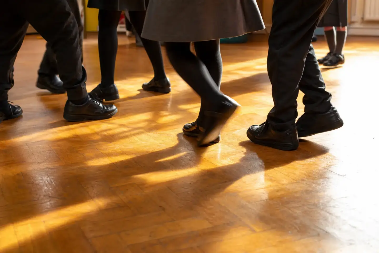 Pupils' feet and lower legs mid-step on a school hall floor, one pair turning on the spot, in warm afternoon light.