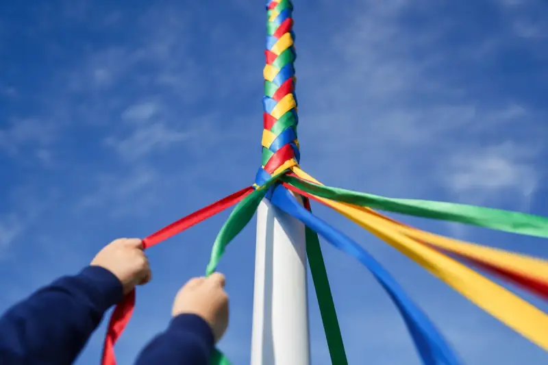 Close view of the top of a maypole where coloured ribbons cross into a woven plait, against a bright overcast sky.